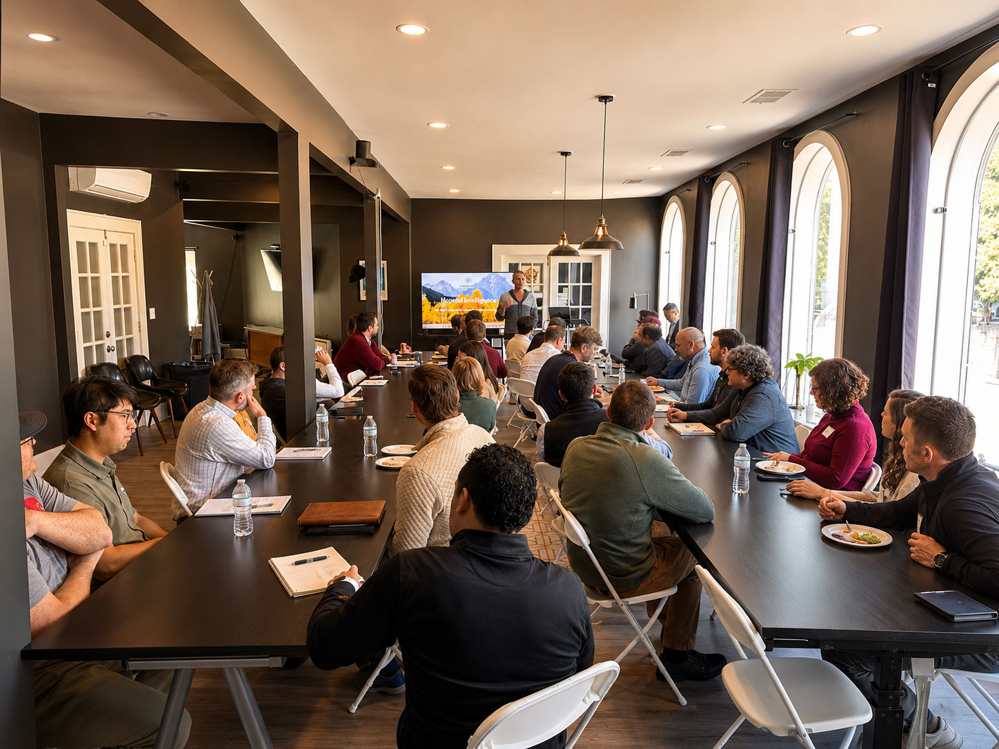 Business leaders seated around long tables at a Triptych leadership session, a presenter speaking beside a screen