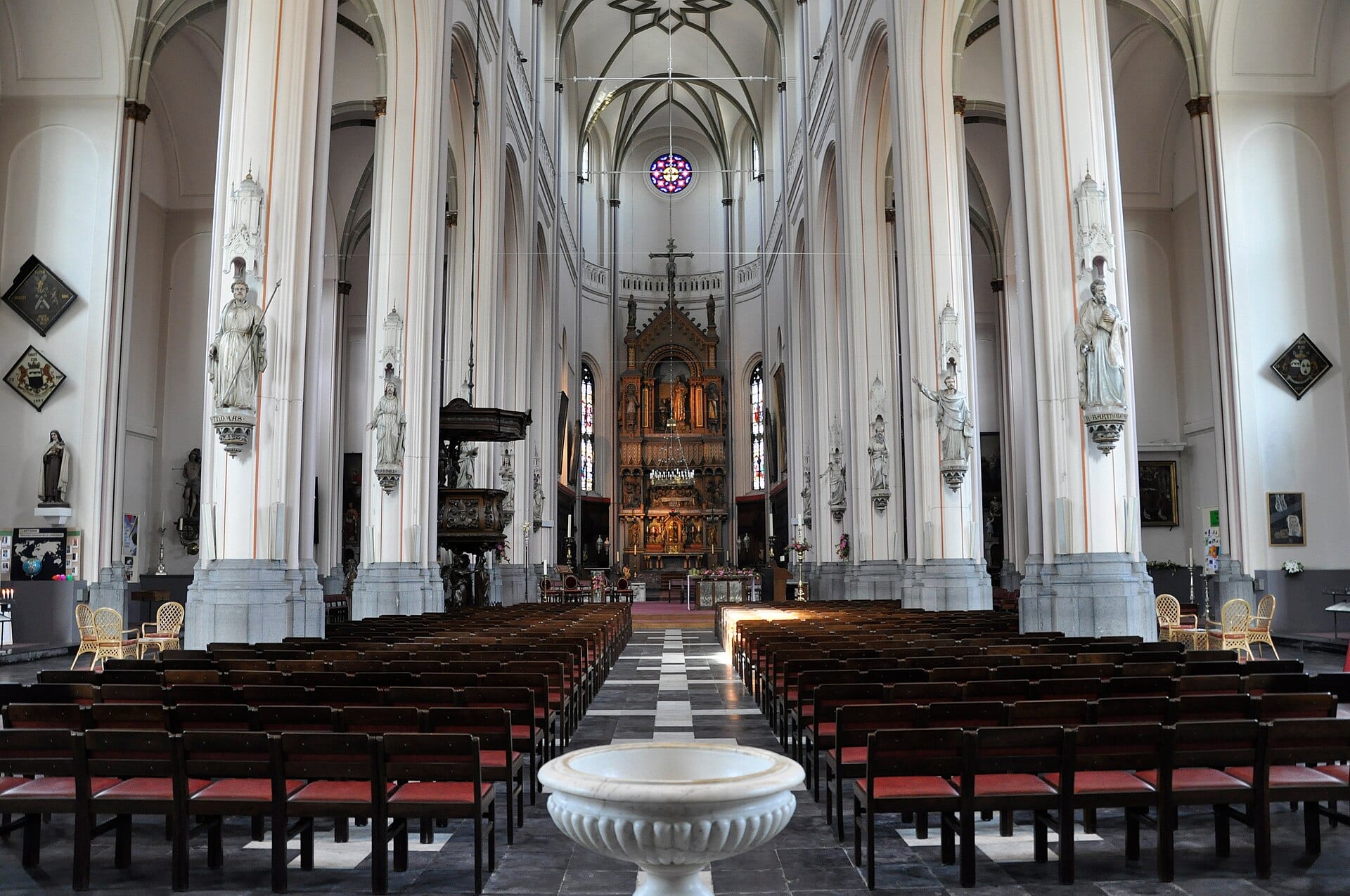 Interior of Sint-Gertrudiskerk in Wetteren, with white columns and rows of chairs facing the high altar.